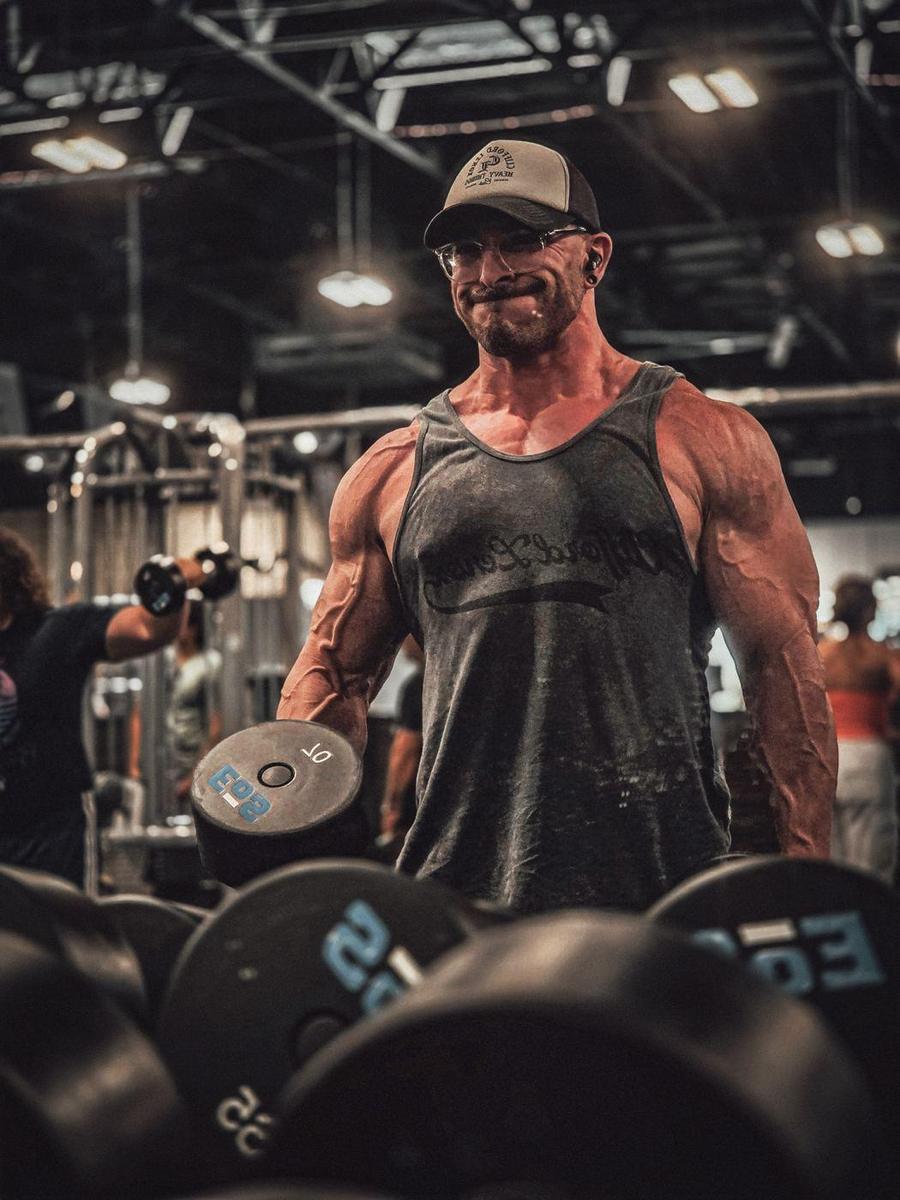 Bodybuilder in a cap and glasses curling a heavy dumbbell in a busy gym