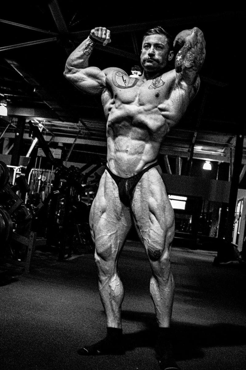 Bodybuilder hitting a front double biceps with one arm behind his head in a dark gym, black and white