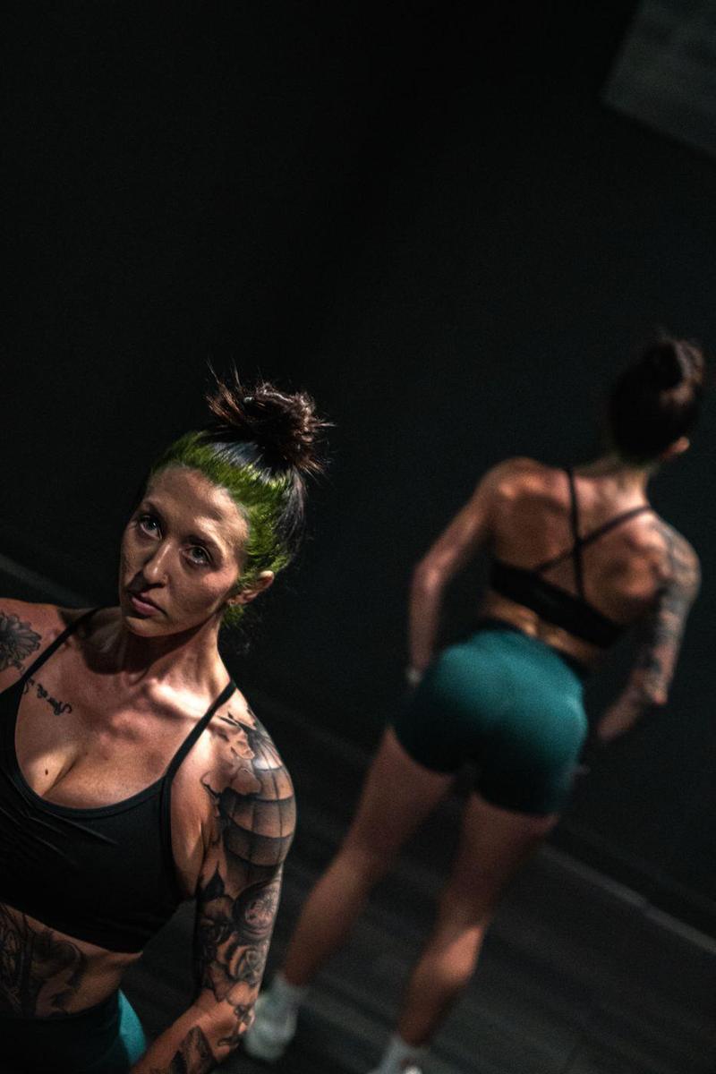 Tattooed athlete with green hair looking up, her reflection behind her in a dark mirror