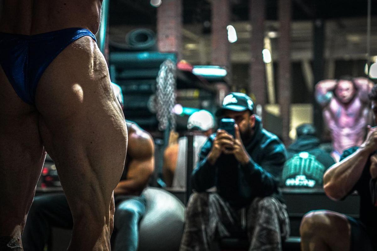 Close-up of a competitor's hamstring during posing practice, people filming in the background